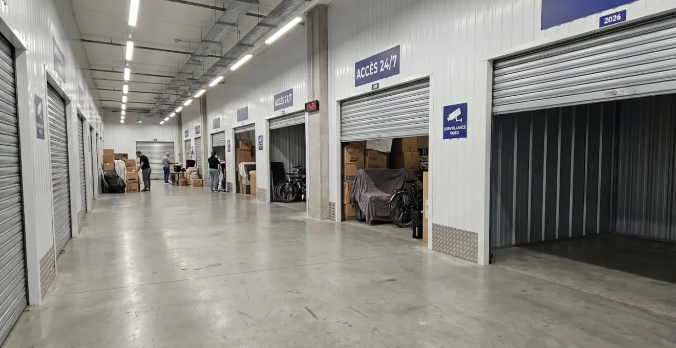 Couloir d'un centre de self-stockage moderne avec plusieurs box de tailles différentes, portes levantes partiellement ouvertes, certains vides et d'autres contenant du mobilier
